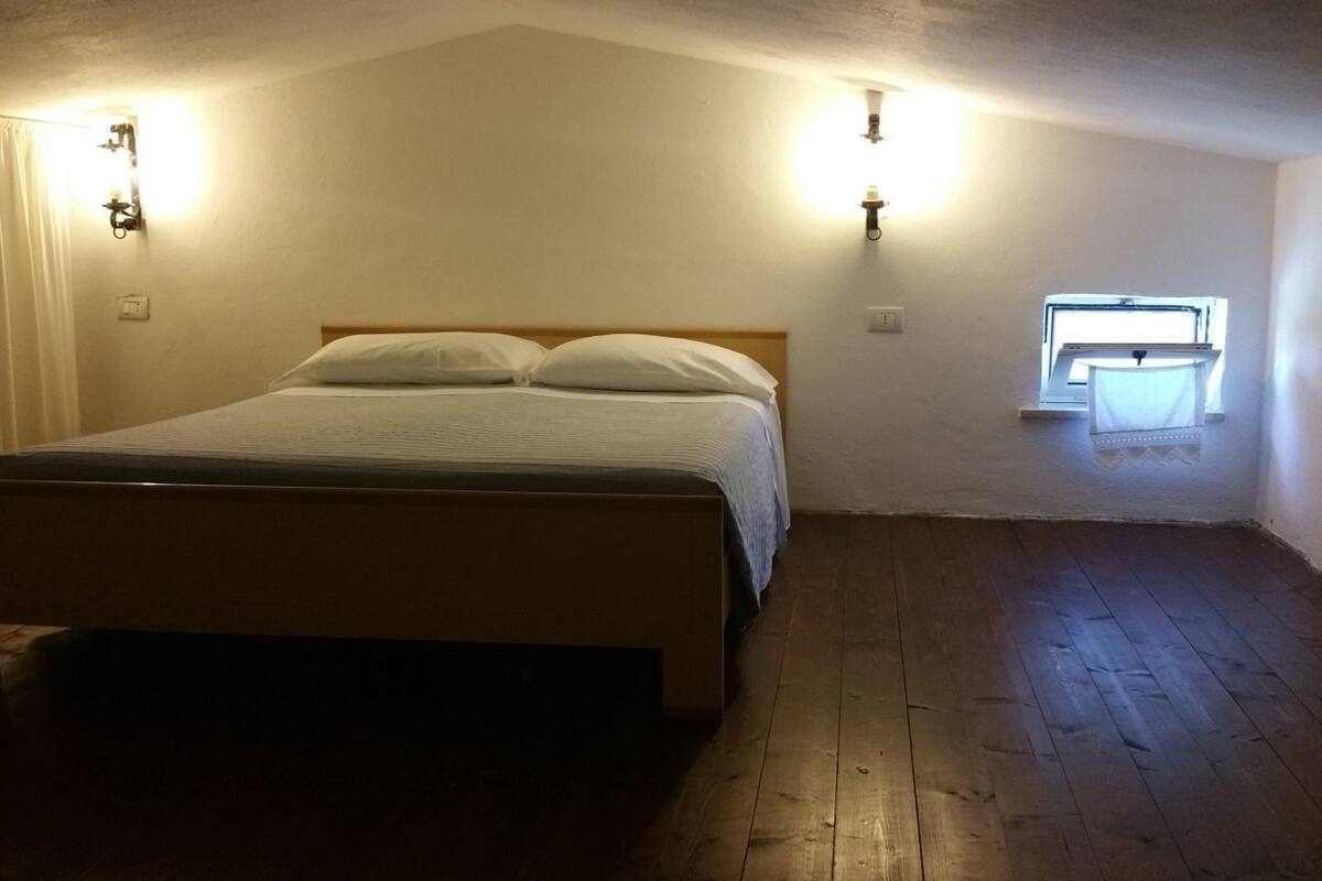 A single bed in a room with wooden flooring, two wall-mounted light fixtures, and a small window with a curtain.