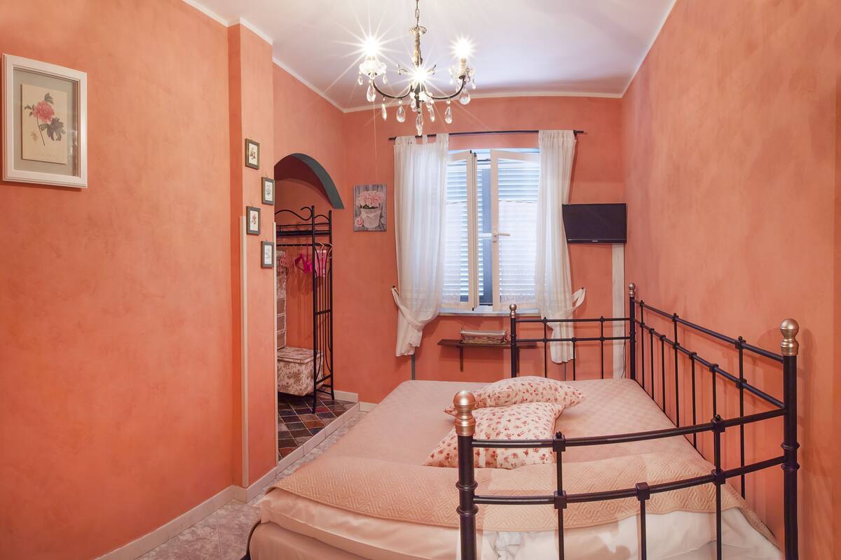 A bedroom with a bed, a chandelier, a window with curtains, and a television.