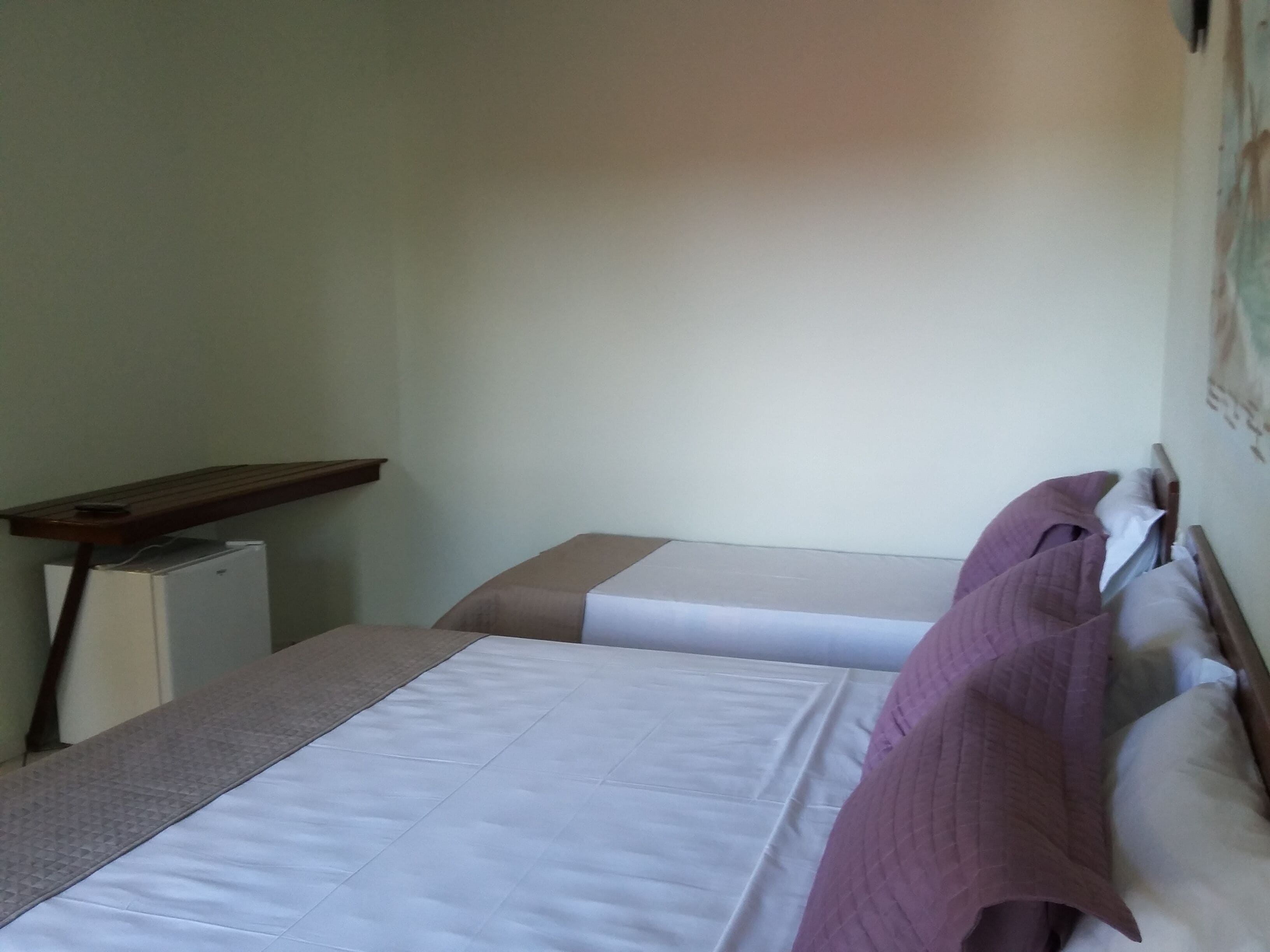 Quarto com duas camas, uma escrivaninha pequena e uma prateleira fixada na parede.