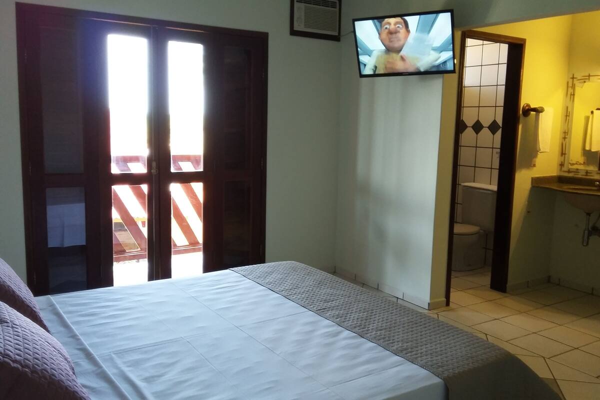 A hotel room with a bed, a television mounted on the wall, a bathroom with a toilet and sink, and a balcony with a wooden railing.