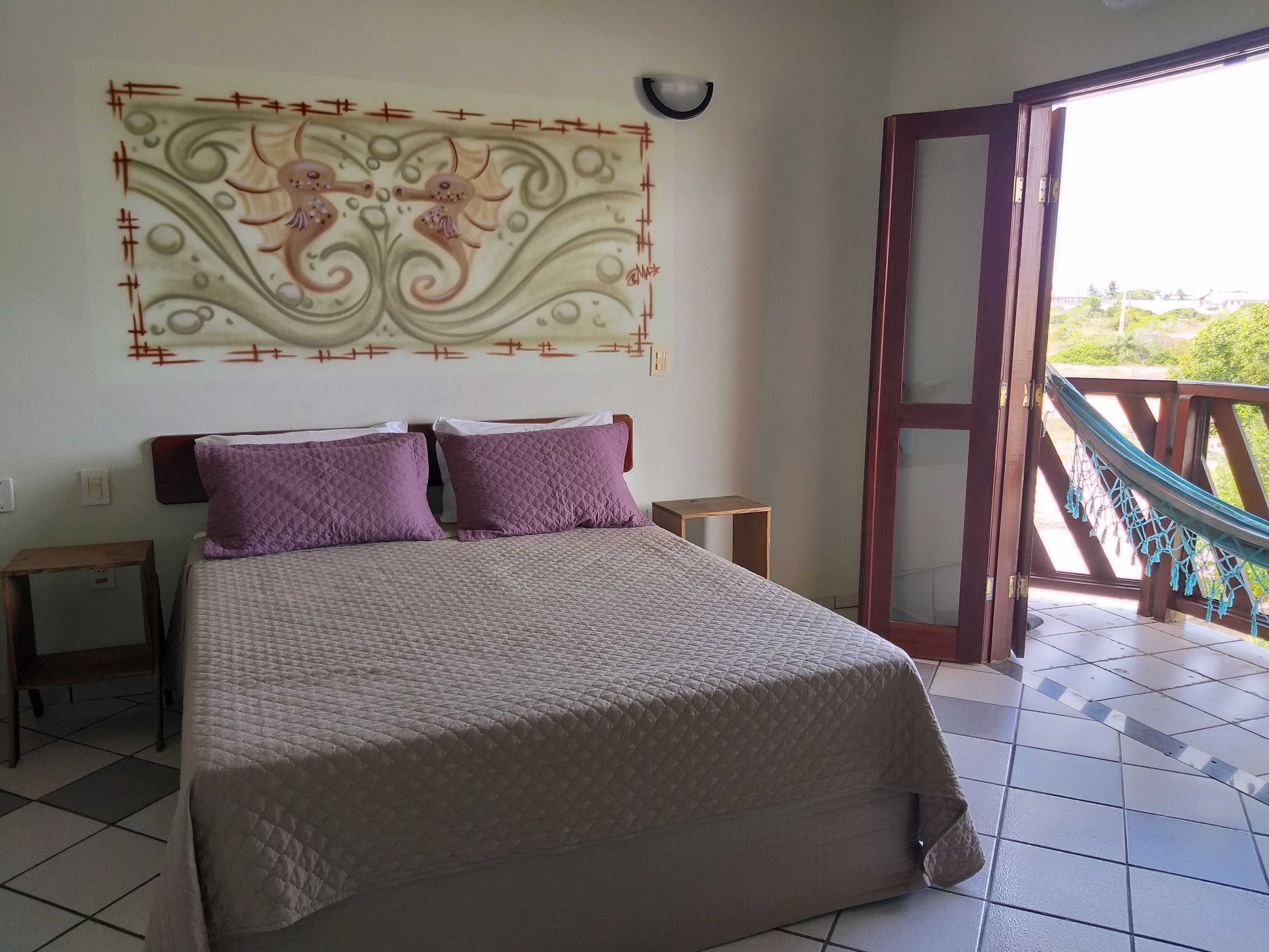 A bed with a quilted cover, two nightstands, a wall-mounted decorative piece, and a view of a balcony with a hammock.