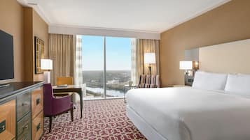 A hotel room with a large bed, a desk with a chair, a TV, and a view of a cityscape through the windows.