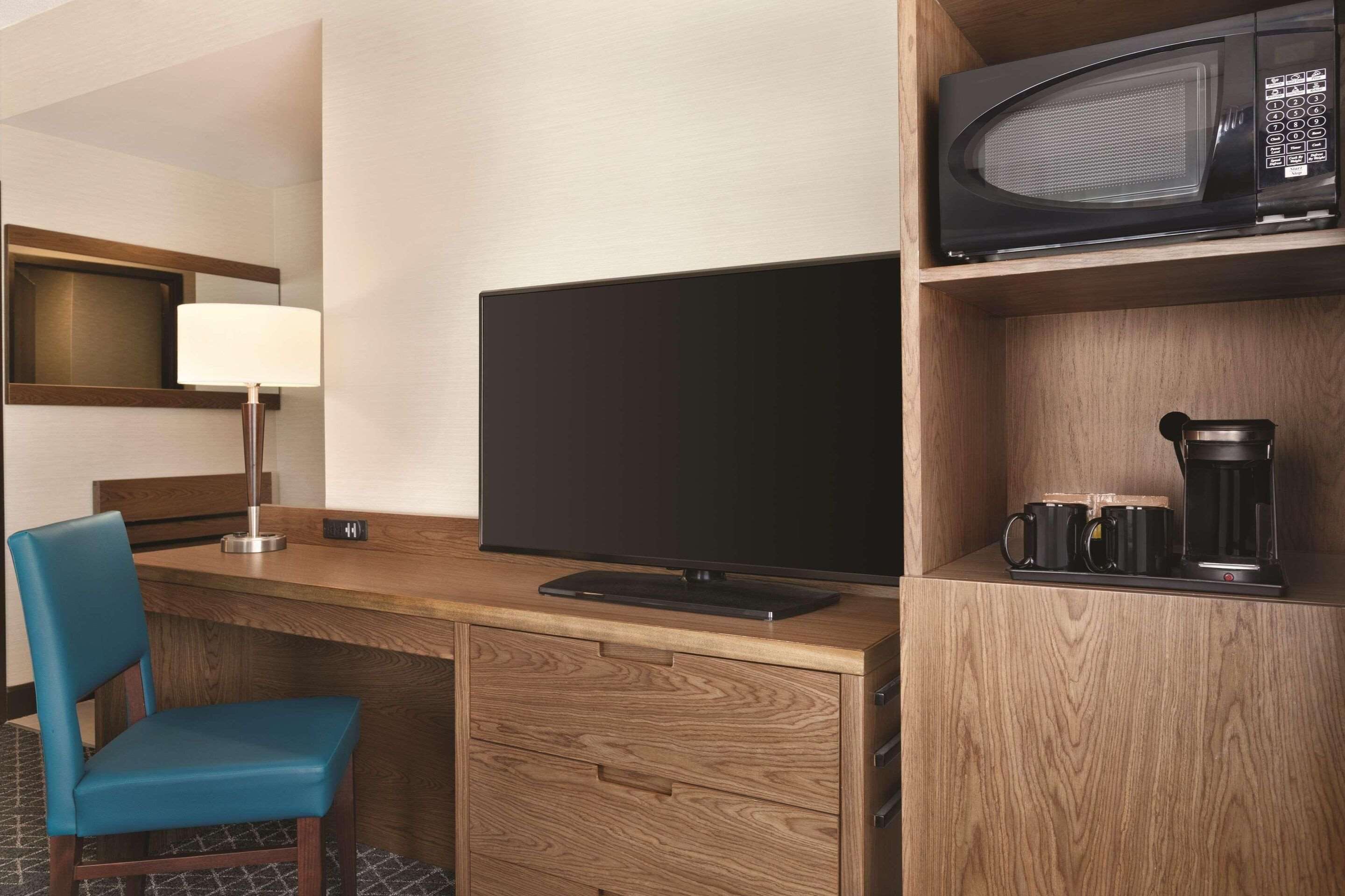 Habitación de hotel con escritorio de madera, televisor de pantalla plana, microondas, cafetera, lámpara y una silla azul.