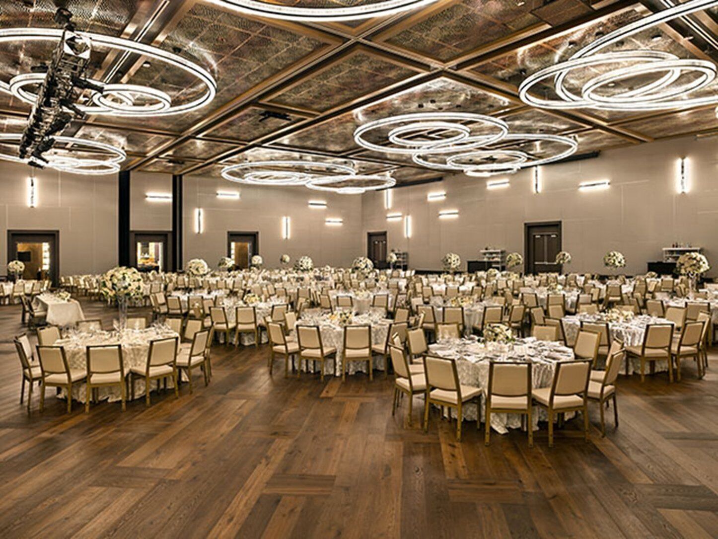 A large banquet hall with round tables and chairs, high ceilings with circular light fixtures, and a polished wooden floor.