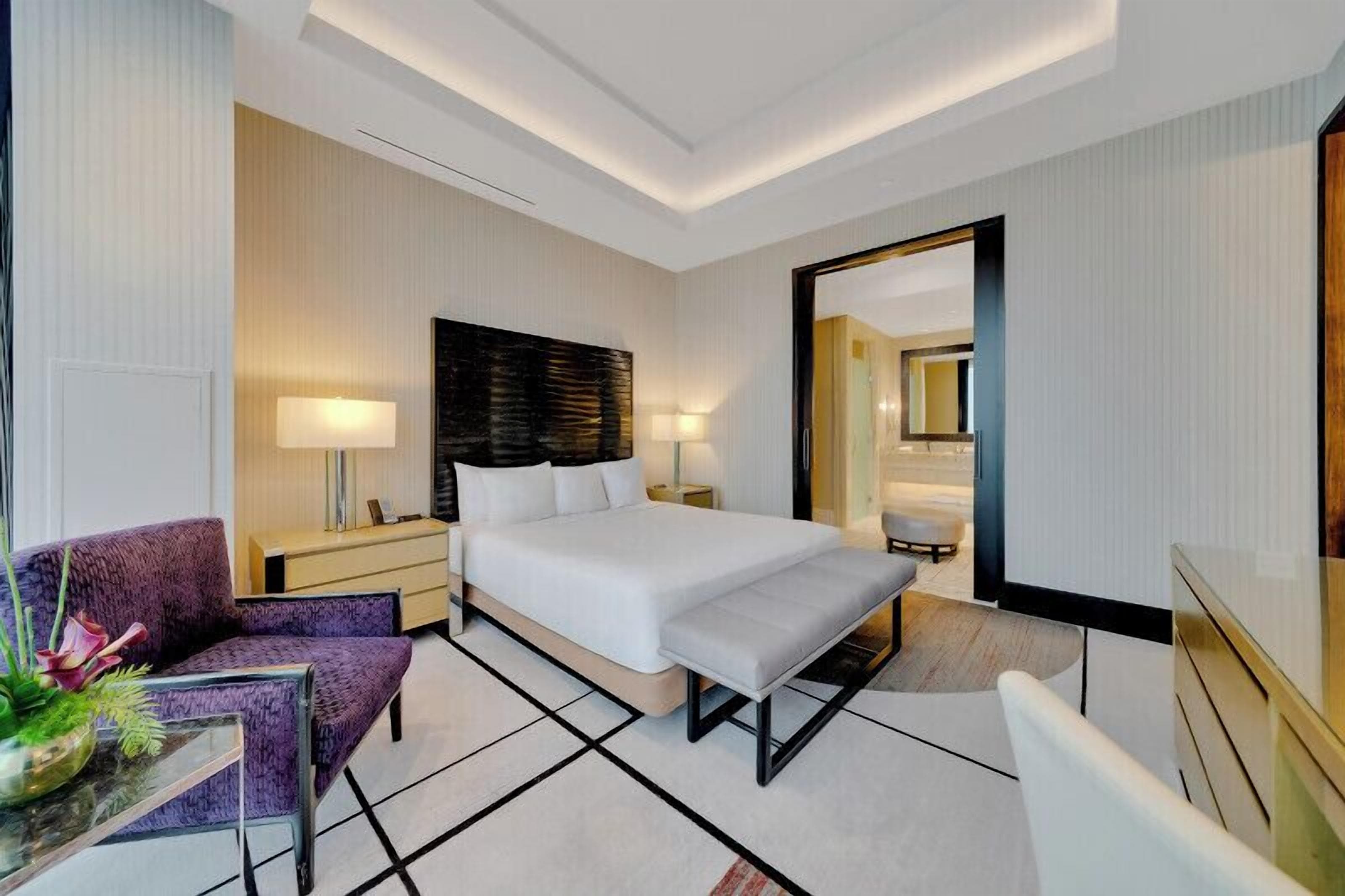 A modern hotel room with a large bed, a desk, a chair, a mirror, and a plant.