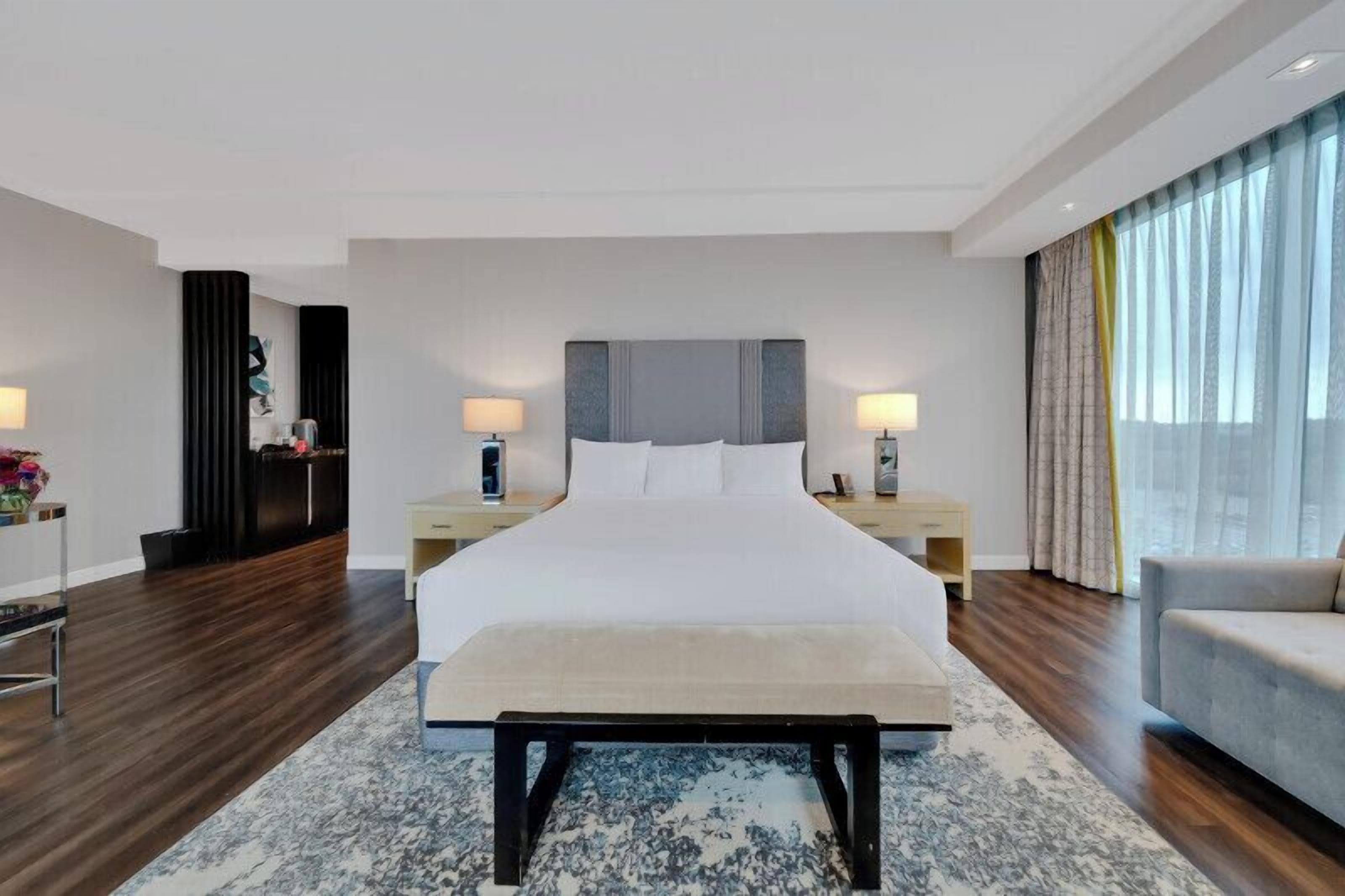 Un hotel moderno con una cama grande, mesitas de noche, un banco y vistas a la ciudad a través de amplias ventanas.