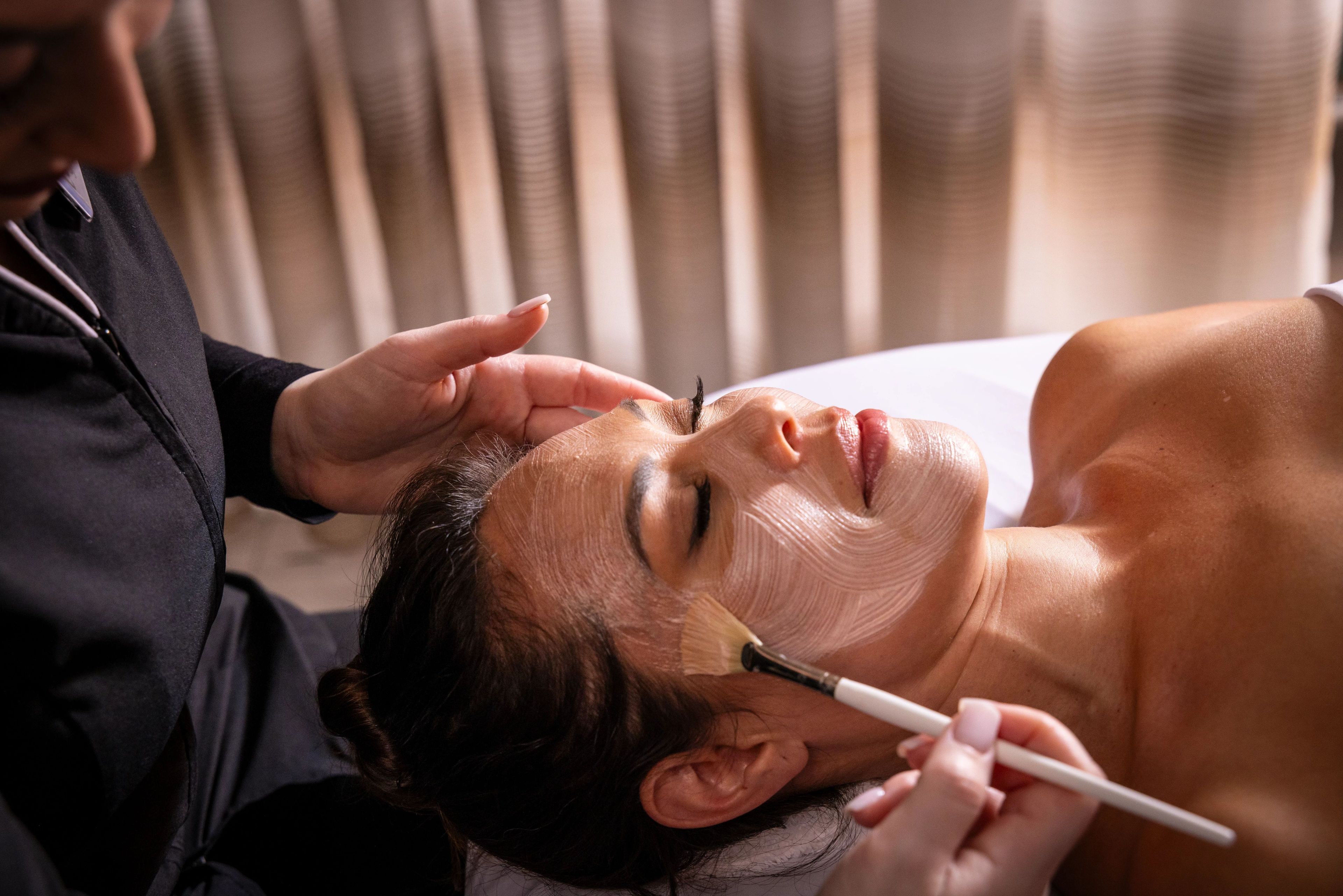 A person lying down with a face mask applied, and another person applying makeup.