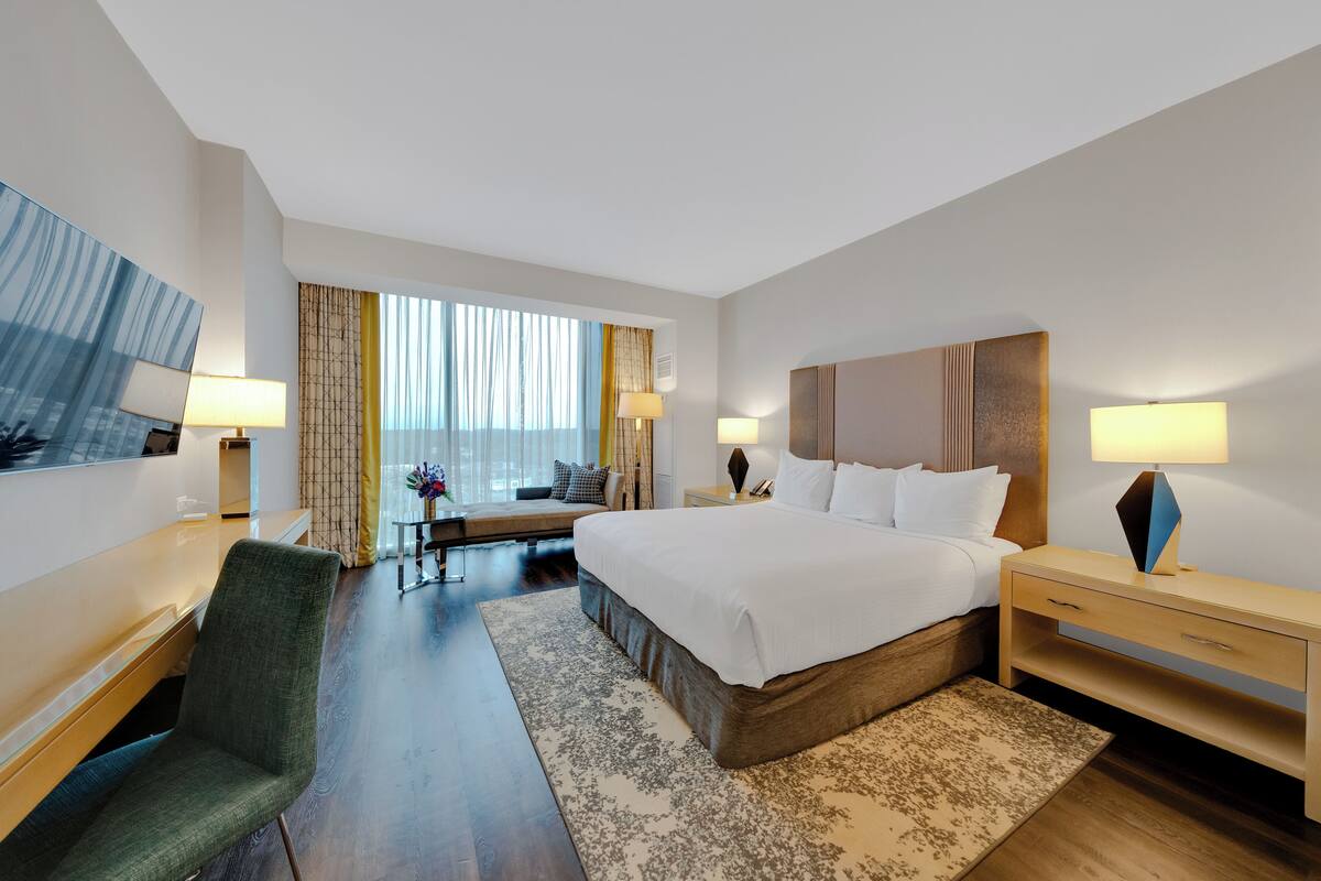 A hotel room with a bed, bedside tables, a desk, a chair, a window with curtains, and a view of the city.