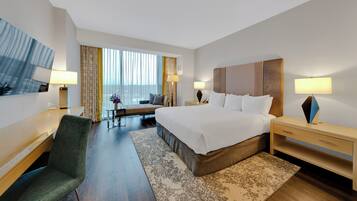 Habitación de hotel con cama, mesitas de noche, escritorio, silla, ventana con cortinas y vistas a la ciudad.