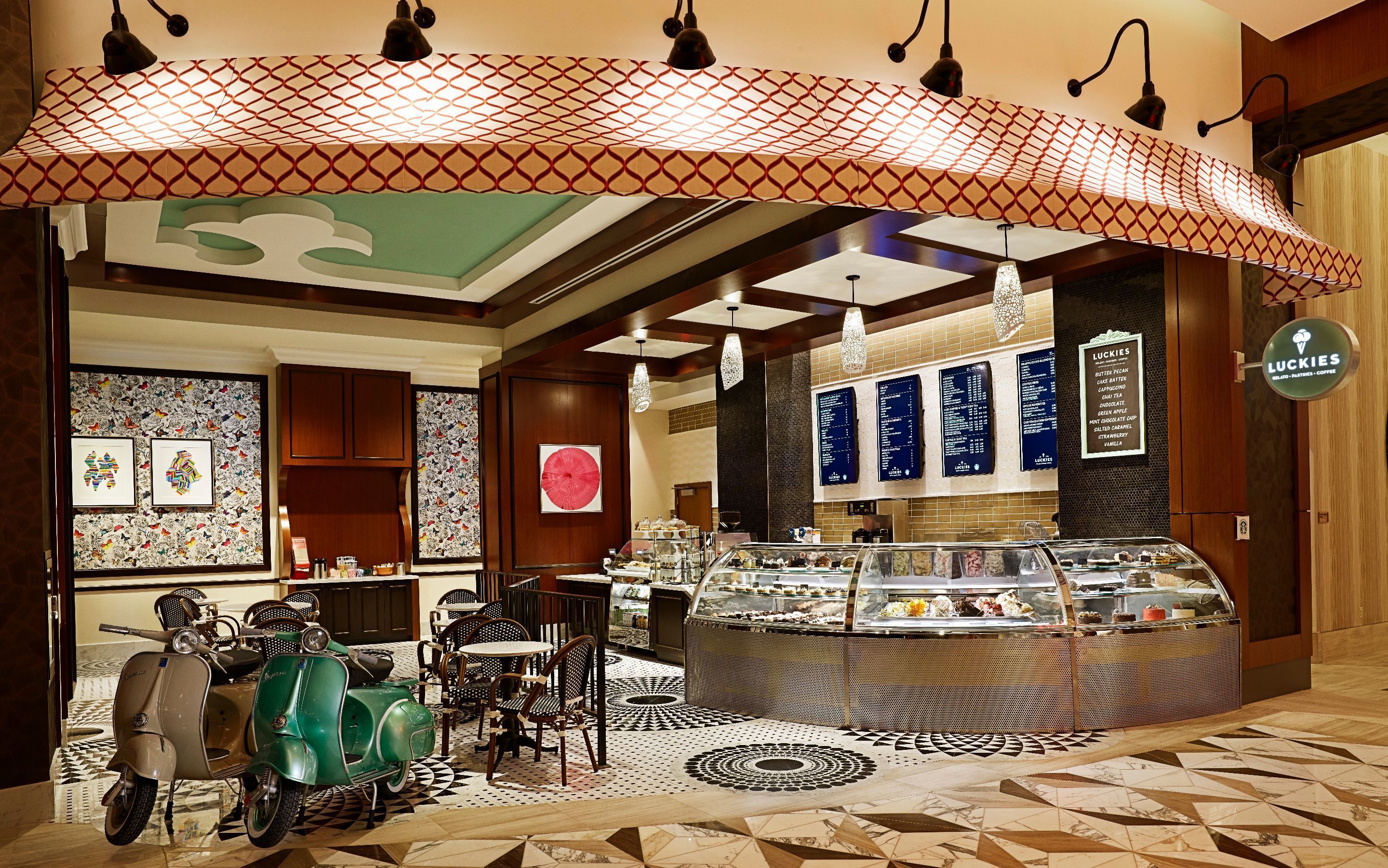 A cafe with a counter displaying various pastries and a menu board. There are chairs and tables arranged for seating.