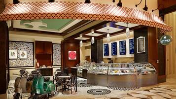 A cafe with a counter displaying various pastries and a menu board. There are chairs and tables arranged for seating.