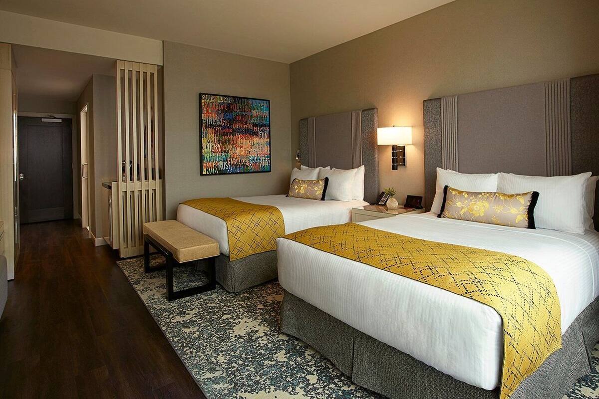 A hotel room with two beds, a bench, a colorful abstract painting, and a nightstand with a lamp.