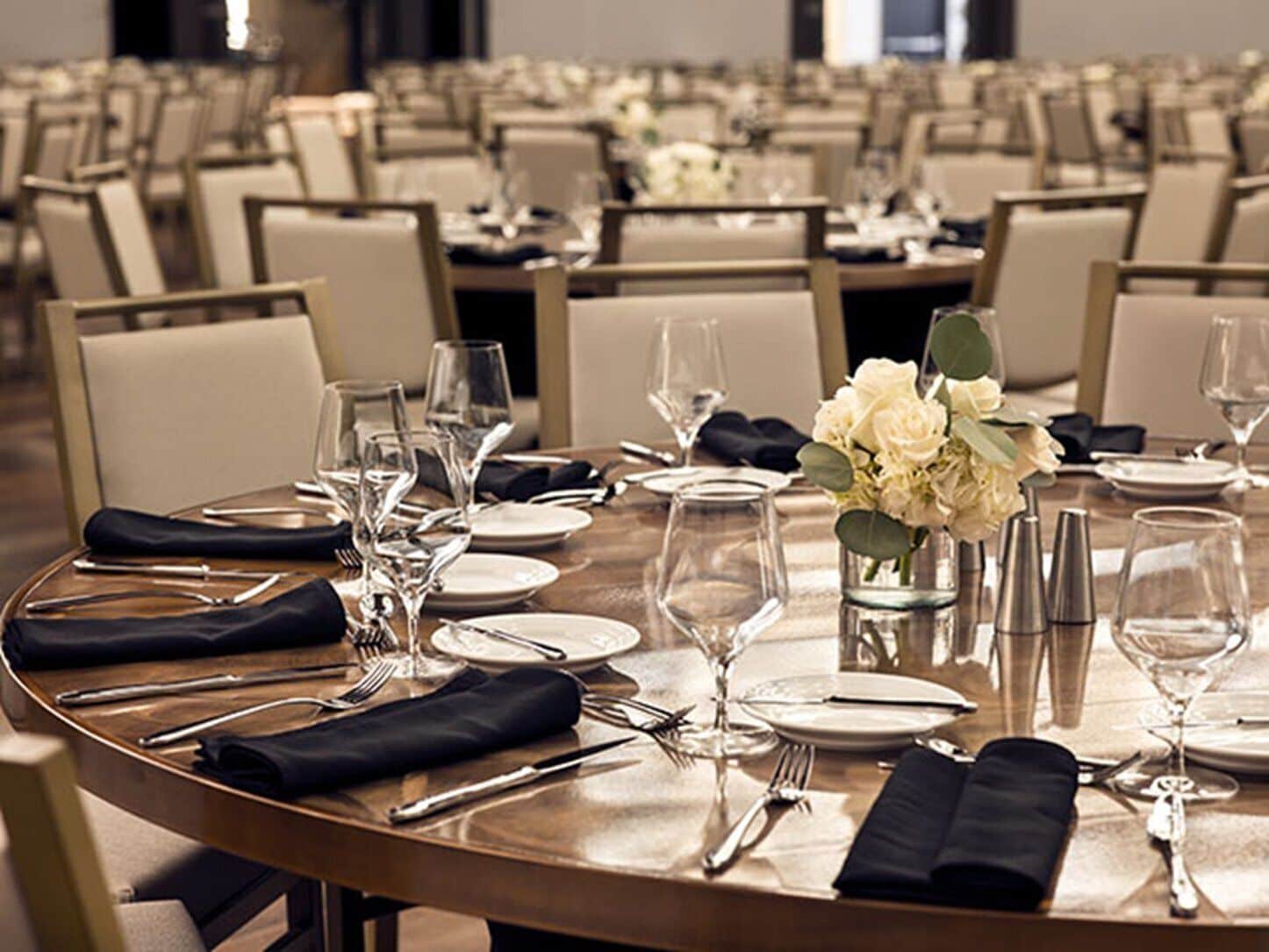 Un salón de conferencias con mesas redondas dispuestas para un evento formal, con sillas, platos, cubiertos y centros de mesa florales.