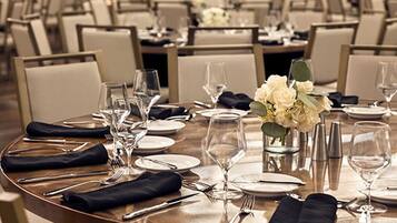 Un salón de conferencias con mesas redondas dispuestas para un evento formal, con sillas, platos, cubiertos y centros de mesa florales.
