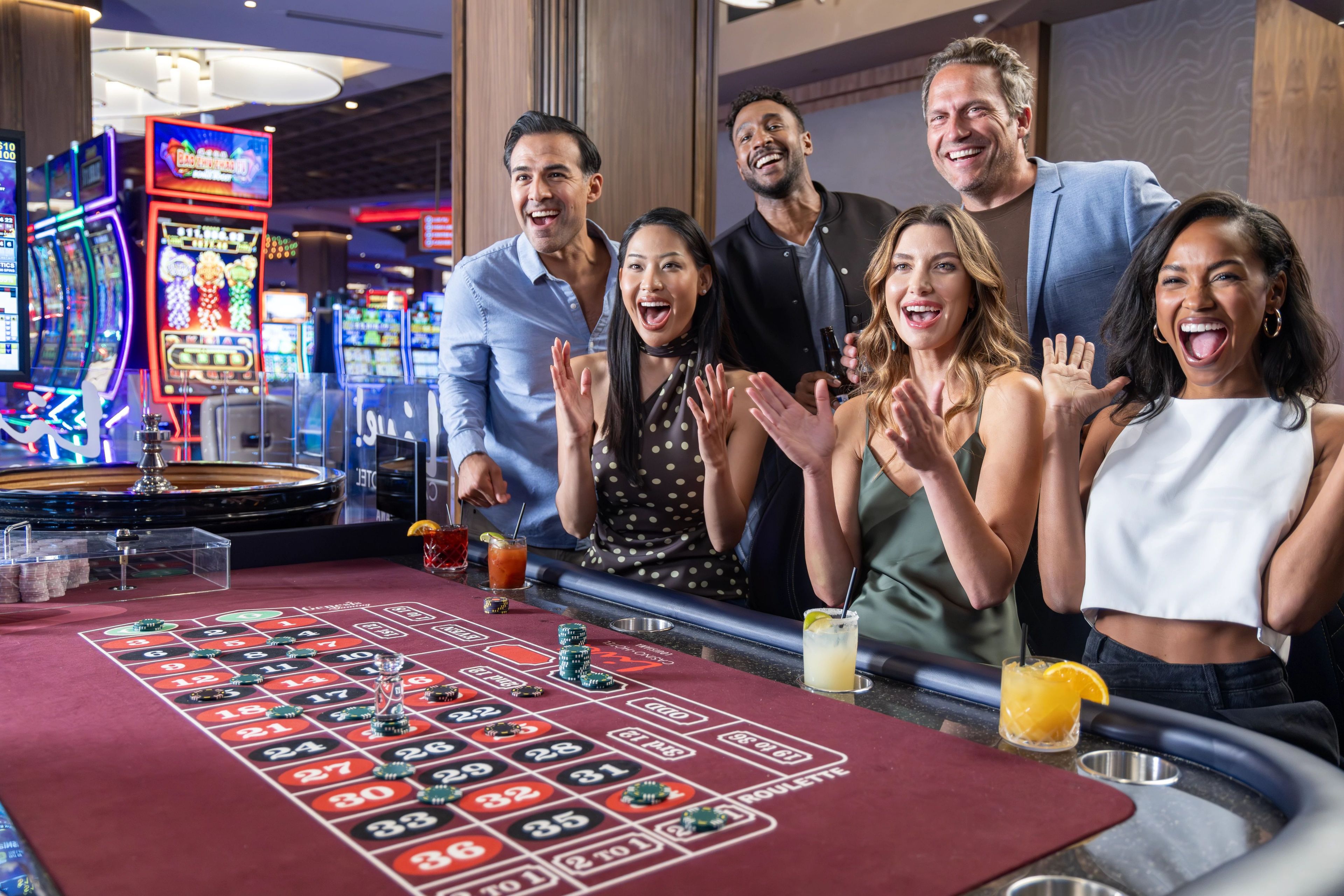 A group of people standing around a roulette table with chips and a drink, in a room with slot machines in the background.