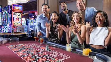 A group of people standing around a roulette table with chips and a drink, in a room with slot machines in the background.
