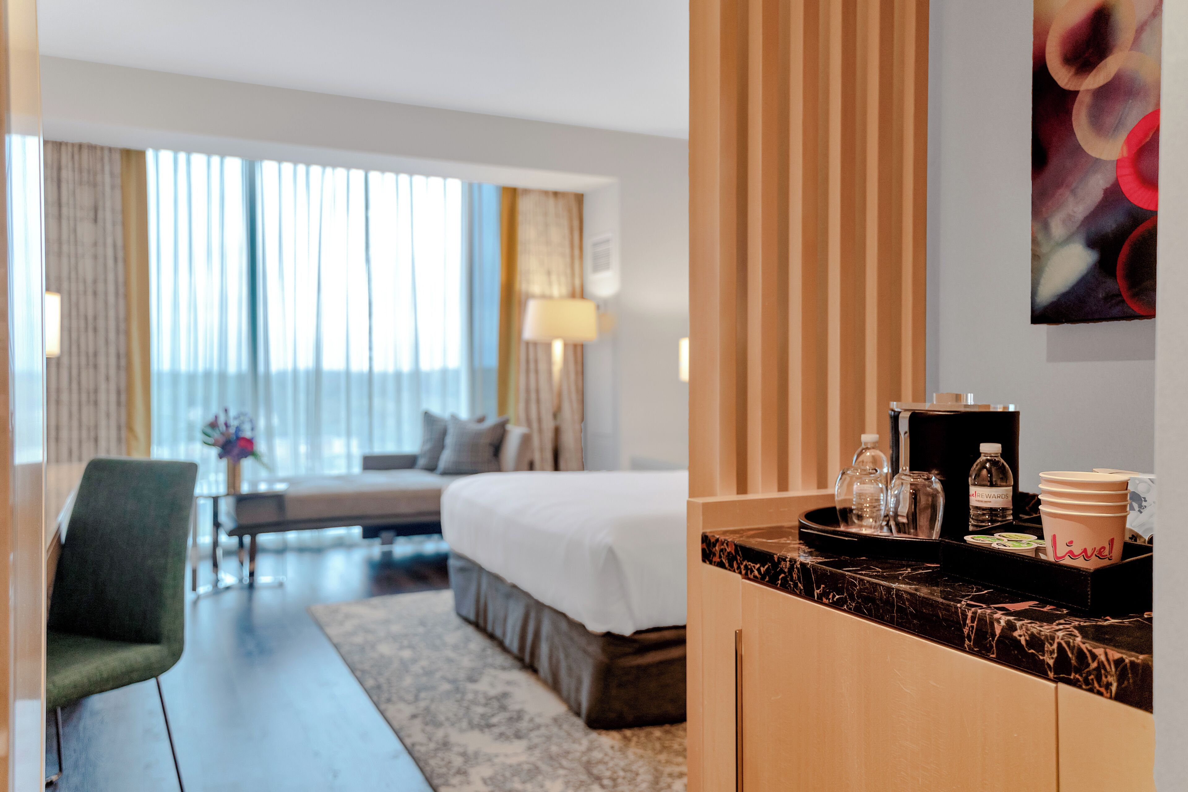 A hotel room with a bed, a desk, a chair, a coffee machine, and a tray with cups and a bottle.