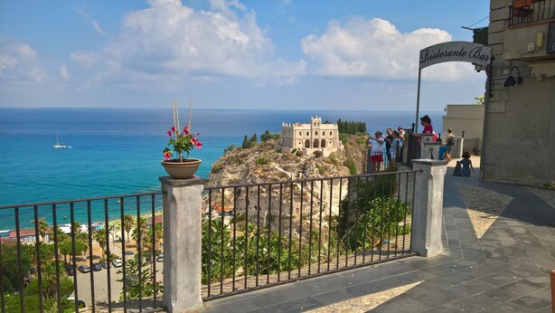 A coastal view with a historic building, a sign for 'Ristorante Bar', and a railing with a potted plant. - Villa Adua (Tropea)