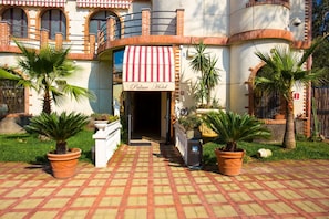 A hotel entrance with a red and white striped awning, a brick facade, and potted palm trees. - Palace Hotel Costa Del Sole (Catania)