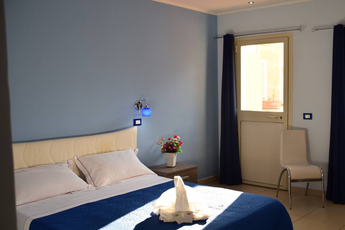 A neatly made bed with a blue bedspread, a bedside table with a vase of flowers, and a chair in a room with light blue walls.