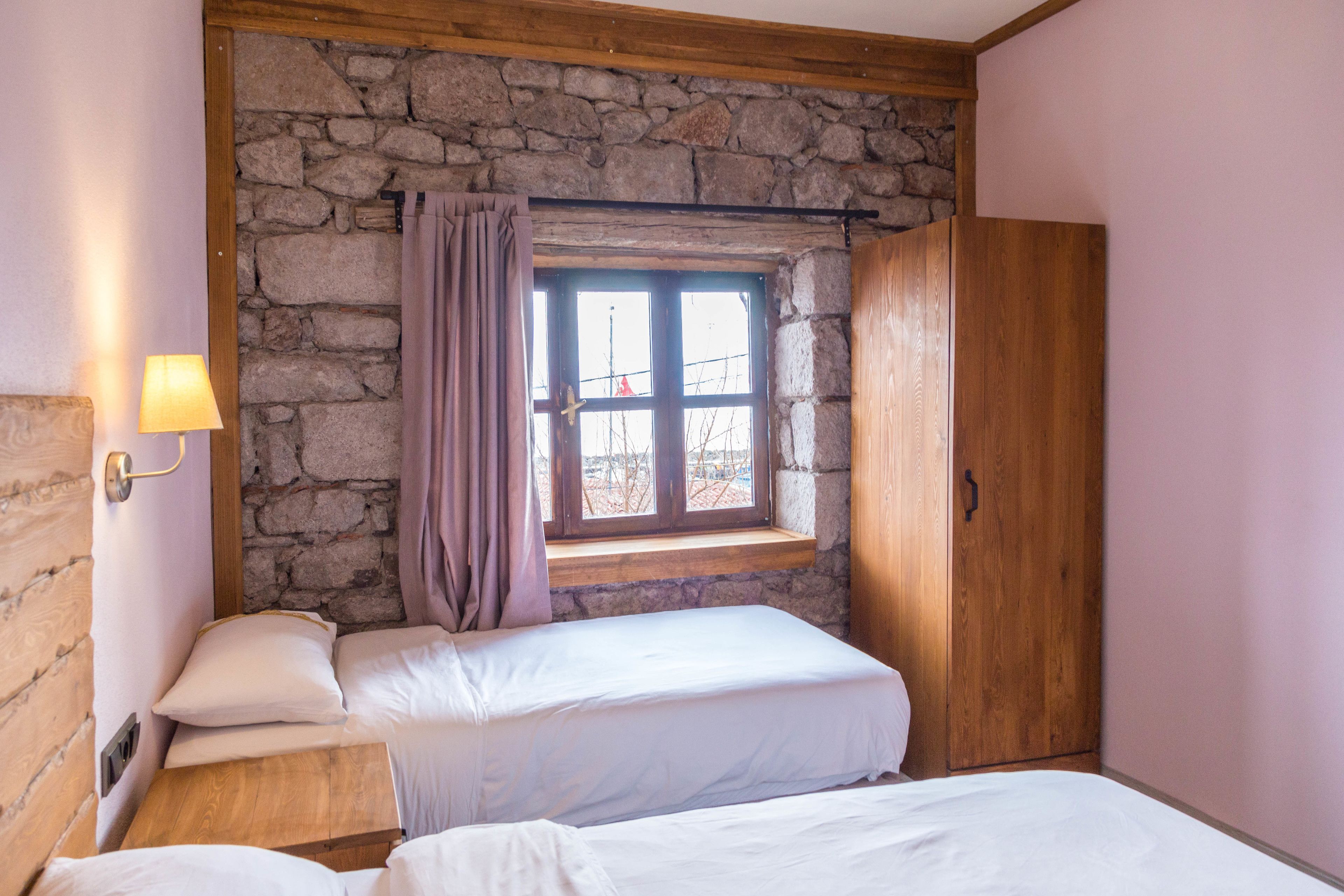 Un dormitorio con dos camas, una pared de piedra, techo de madera, una ventana con cortinas y un armario de madera.
