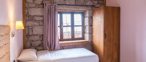 A bedroom with two beds, a stone wall, a wooden ceiling, a window with curtains, and a wooden wardrobe.