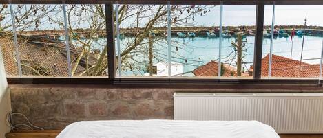 A room with a bed, a view of boats on water, and a stone wall.