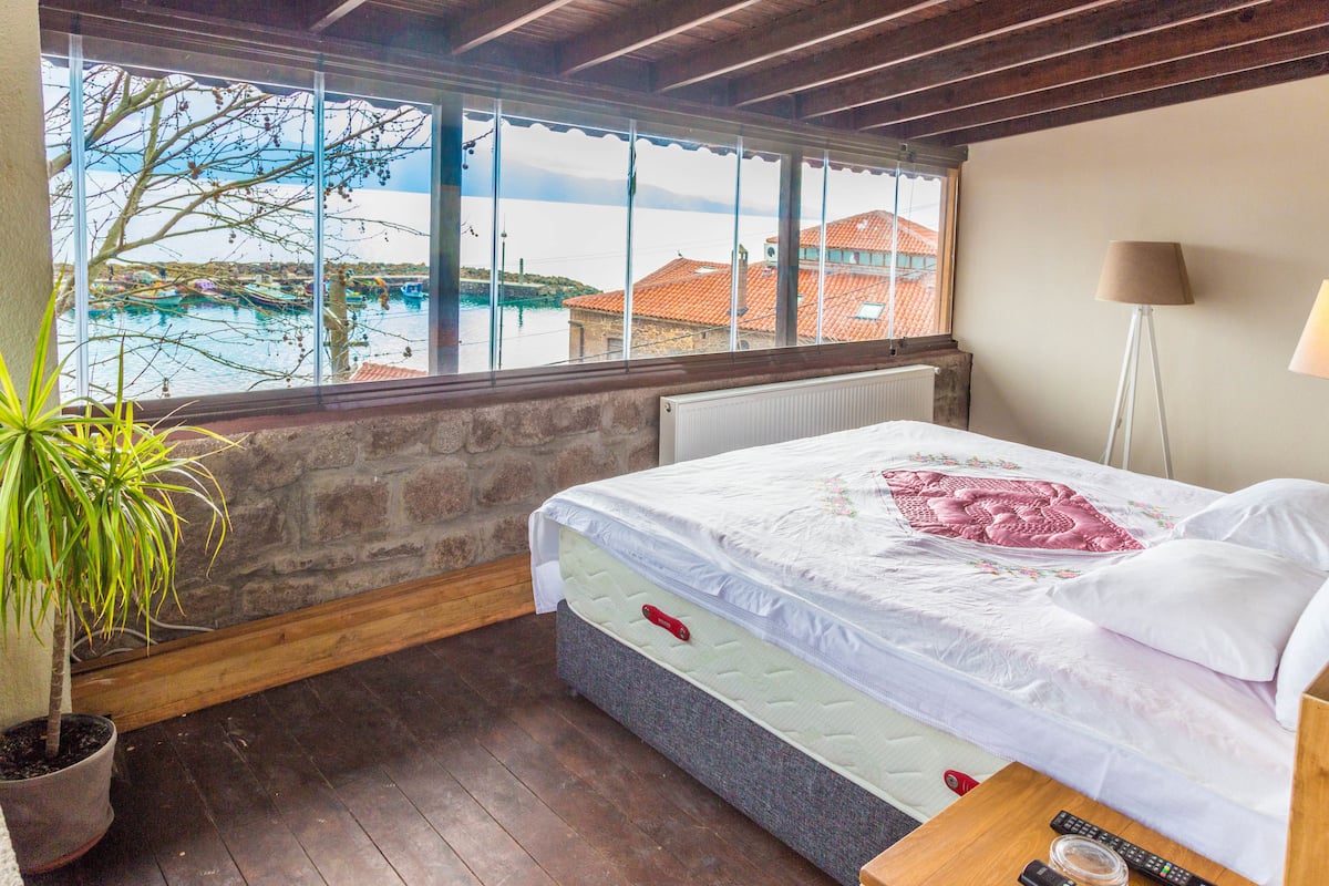 Quarto com uma janela grande, uma cama, mesa de cabeceira, abajur e uma planta em vaso.