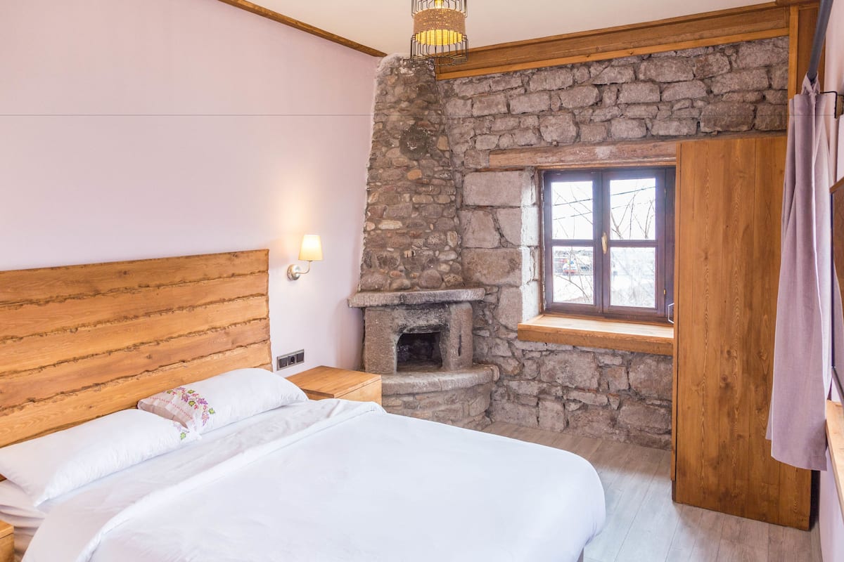 Um quarto com cabeceira de madeira, lareira de pedra, uma janela e mesa de cabeceira de madeira.