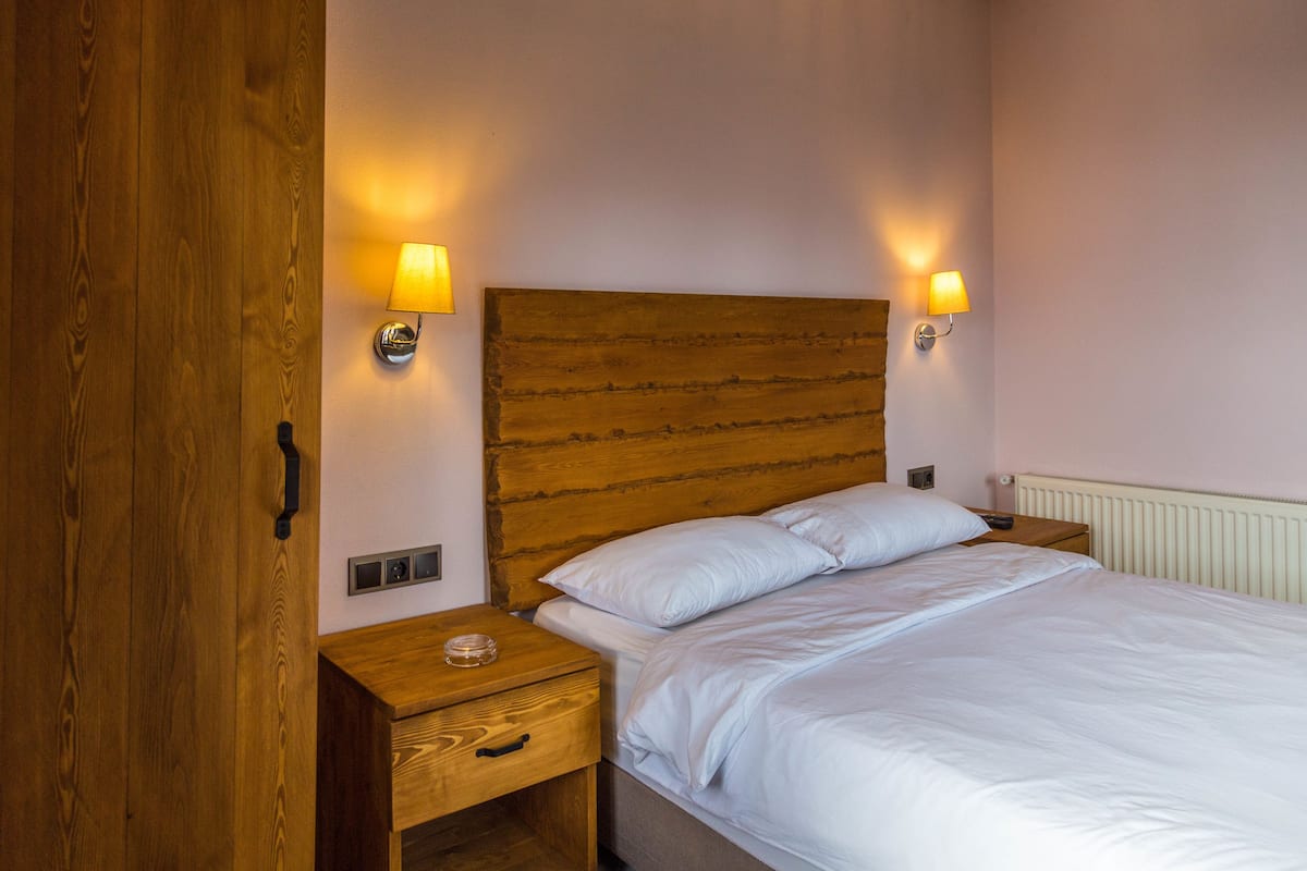 Quarto com cabeceira de madeira, duas luminárias de apoio, mesa de cabeceira e cama com roupa de cama branca.