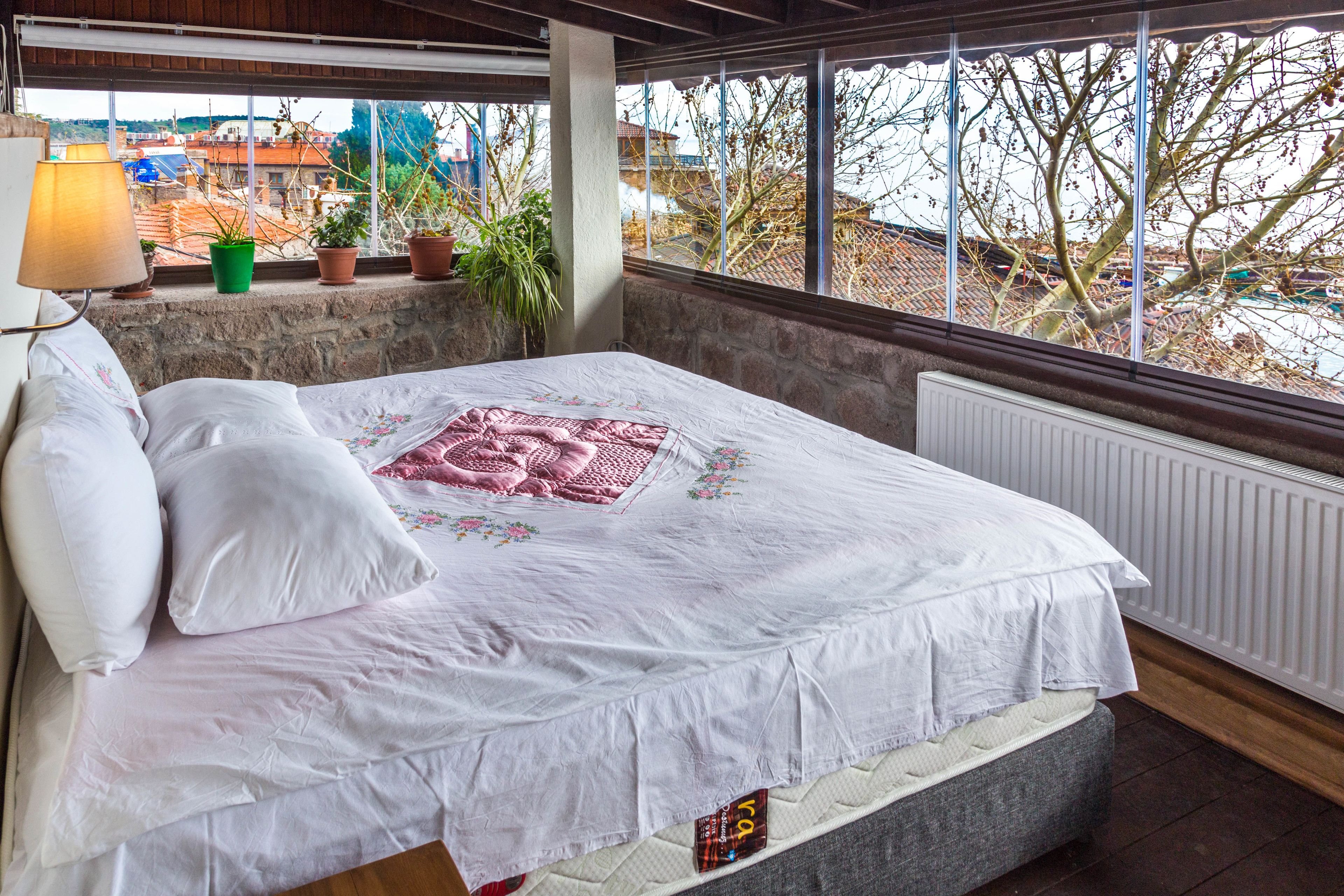 A bedroom with a bed, pillows, a radiator, a window with a view of buildings and trees, and a lamp.