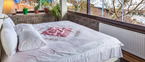 Un dormitorio con cama, almohadas, un radiador, una ventana con vistas a edificios y árboles, y una lámpara.