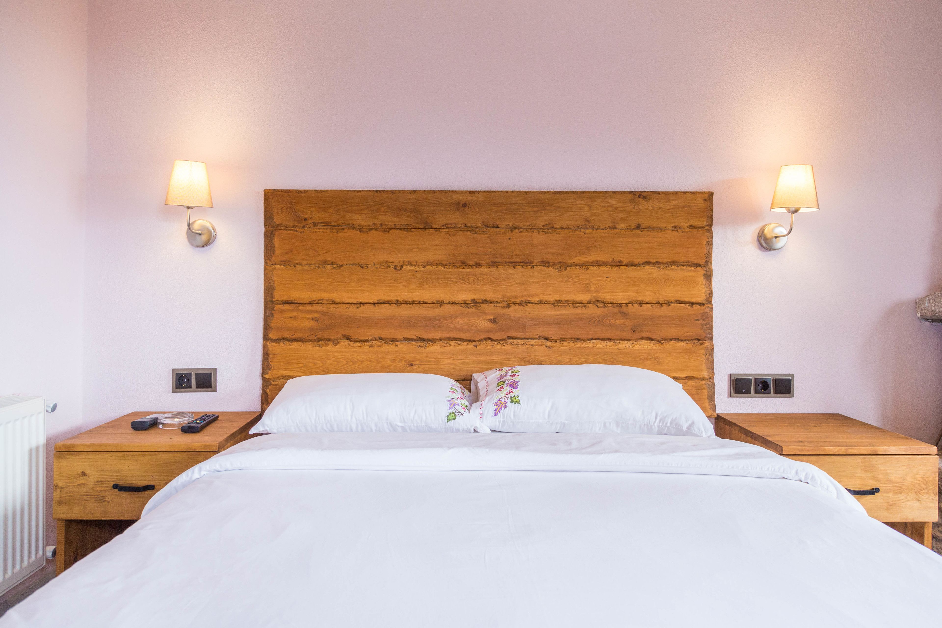 Una cama bien hecha con sábanas blancas y almohadas estampadas, flanqueada por mesitas de noche de madera con lámparas, en una habitación con cabecera de madera y paredes de tonos claros.