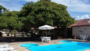 Una piscina con sillas de descanso y una mesa, rodeada de árboles y un edificio con techo de tejas.