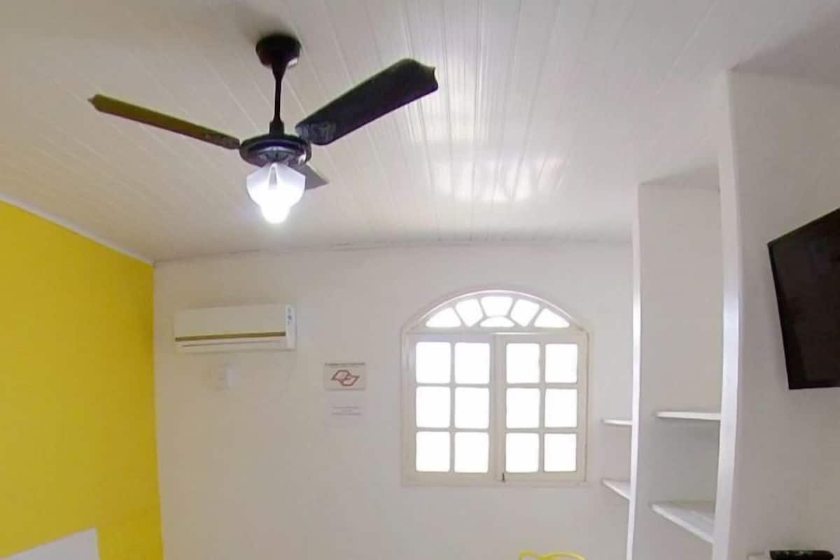 Una habitación con cama, una silla, un televisor, un ventanal y un ventilador de techo.