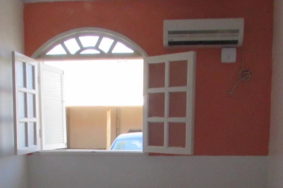 Una cama individual con ropa blanca y almohadas, una mesita de noche pequeña y una ventana con persiana abierta.