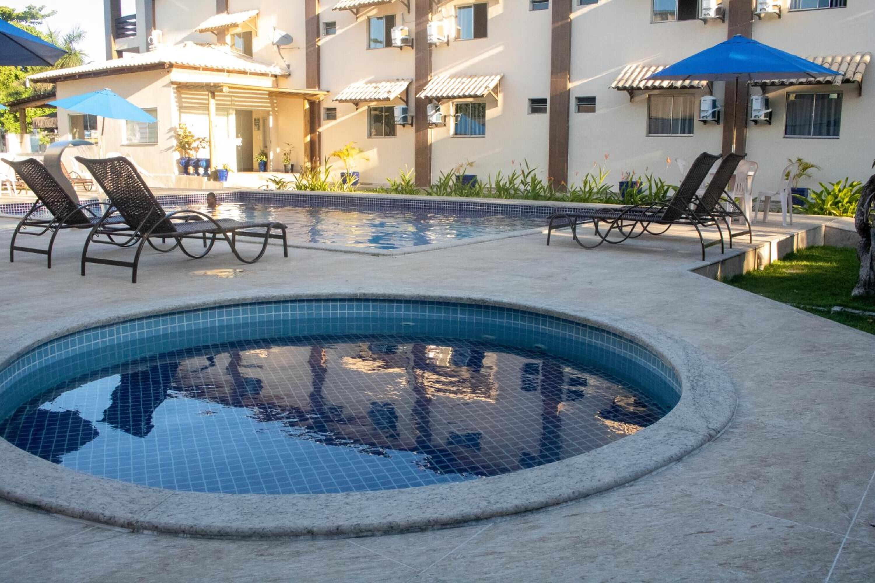 A swimming pool with lounge chairs and umbrellas, surrounded by a building with multiple balconies and air conditioning units.