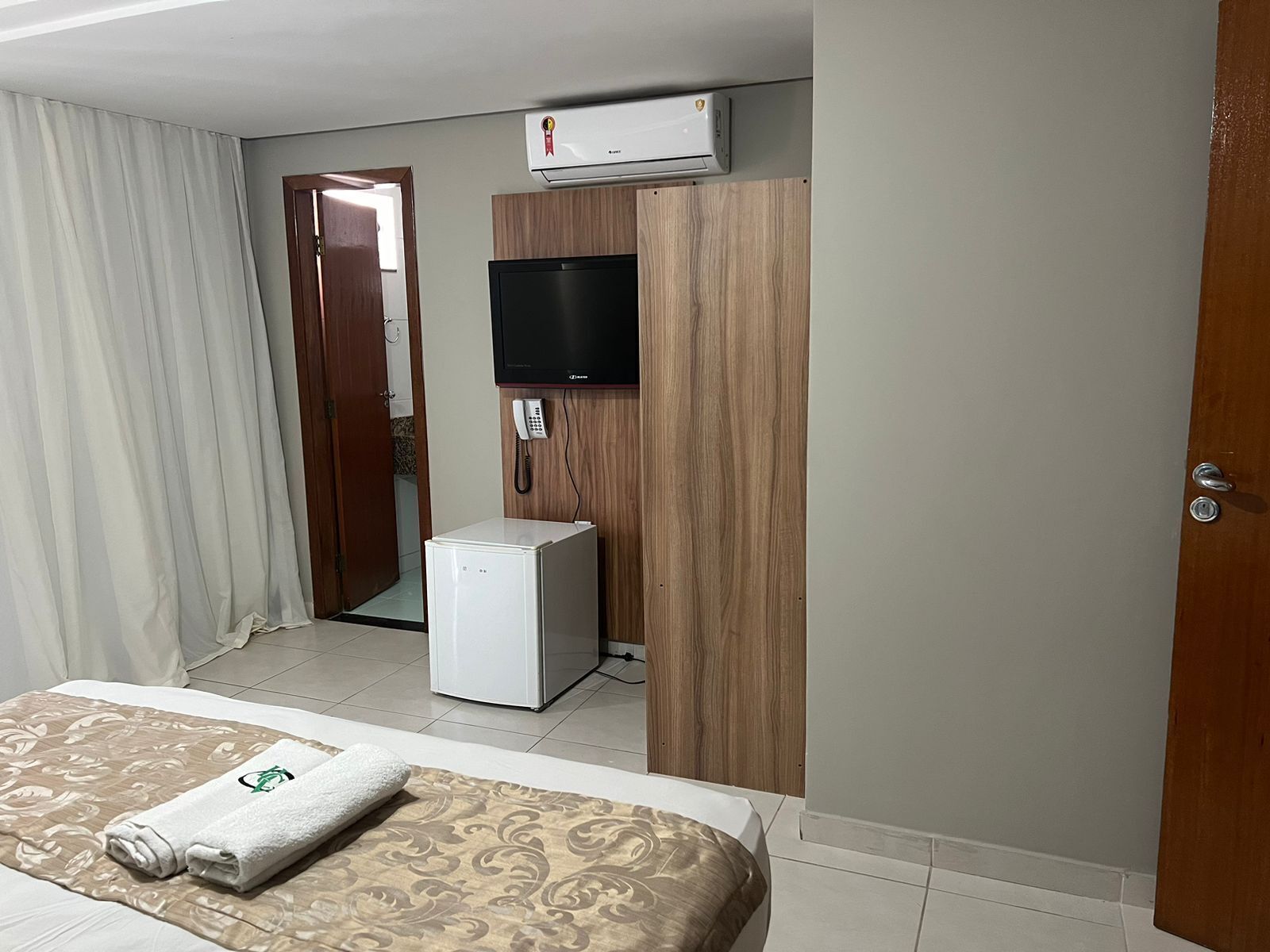 A hotel room with a bed, a small refrigerator, a telephone, and an air conditioning unit.