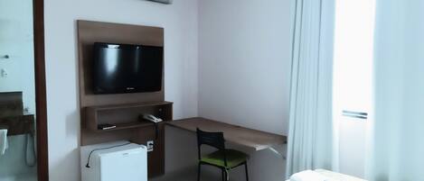 Habitación de hotel con cama, escritorio con silla, televisor, refrigerador pequeño y equipo de aire acondicionado.