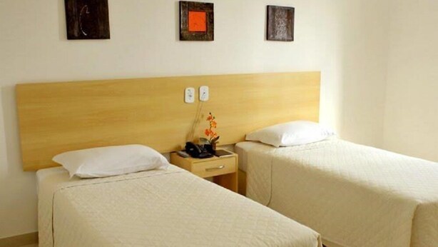 A hotel room with two single beds, a wooden headboard, a nightstand with a phone and a vase of flowers, and three wall-mounted artworks. - Pioneiro Hotel (Teixeira de Freitas)