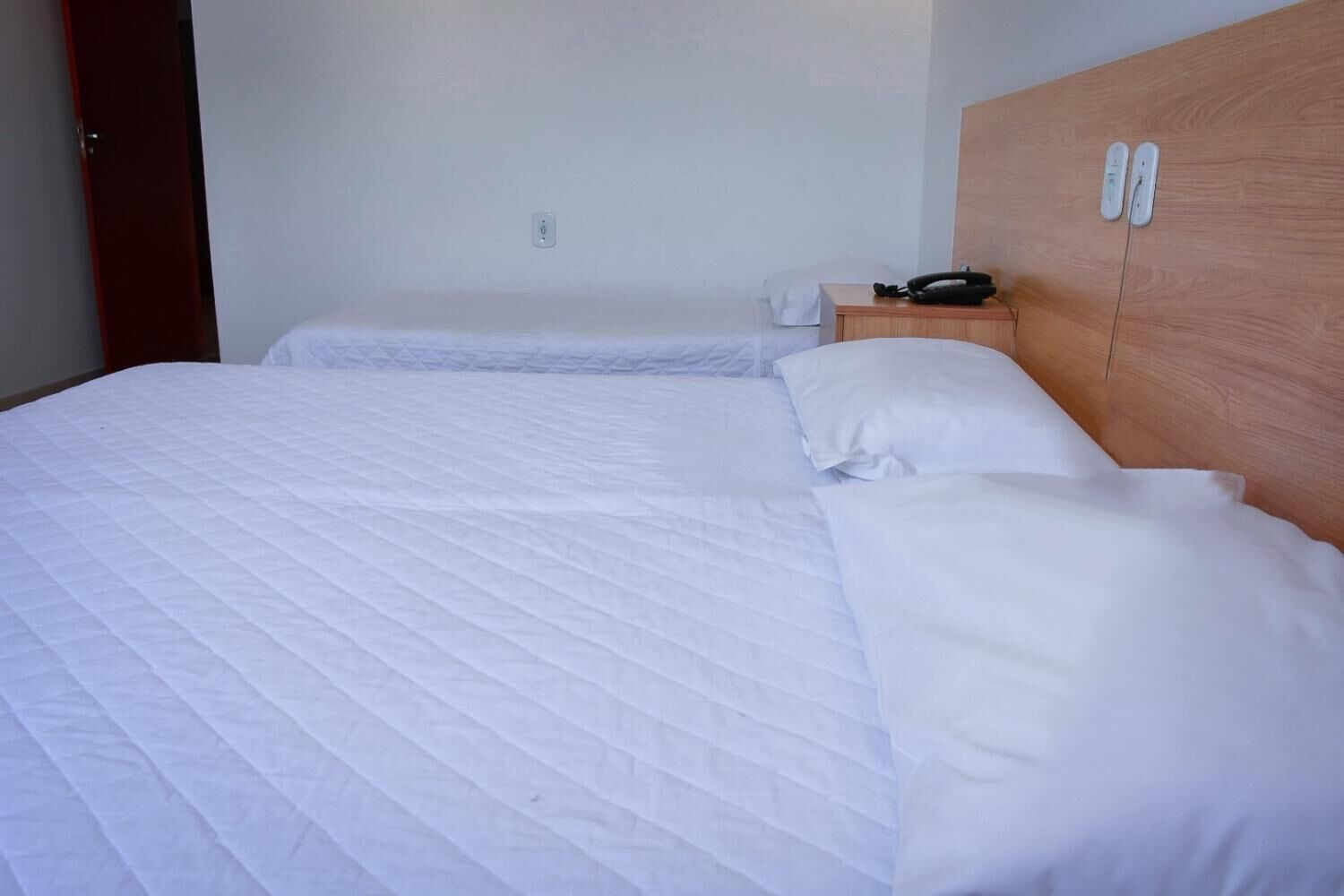 A hotel room with a large bed, a wooden headboard, a bedside table with a phone, and a wall socket.