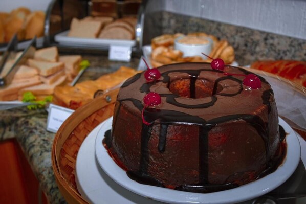 A chocolate cake with cherries on a plate, surrounded by various pastries and bread.