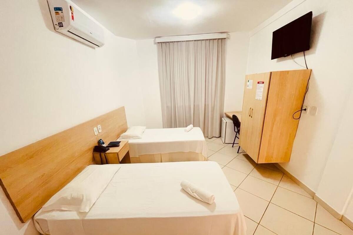 A hotel room with two beds, a wardrobe, a desk, a chair, a TV, and an air conditioning unit.