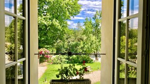 Une vue à travers une fenêtre ornée d’un portail en fer forgé, donnant sur un jardin avec une pelouse et un banc.