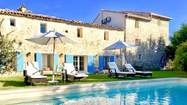Un espace aménagé au bord de la piscine, avec des chaises longues et des parasols, un bâtiment en pierre aux volets bleus et une piscine à l’eau cristalline.