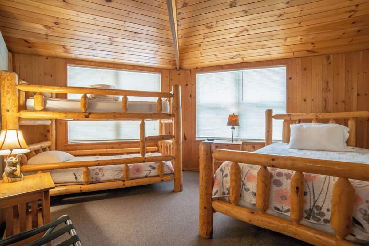 A cabin with wooden walls and floors, a bunk bed, a desk with a lamp, and a window with blinds.