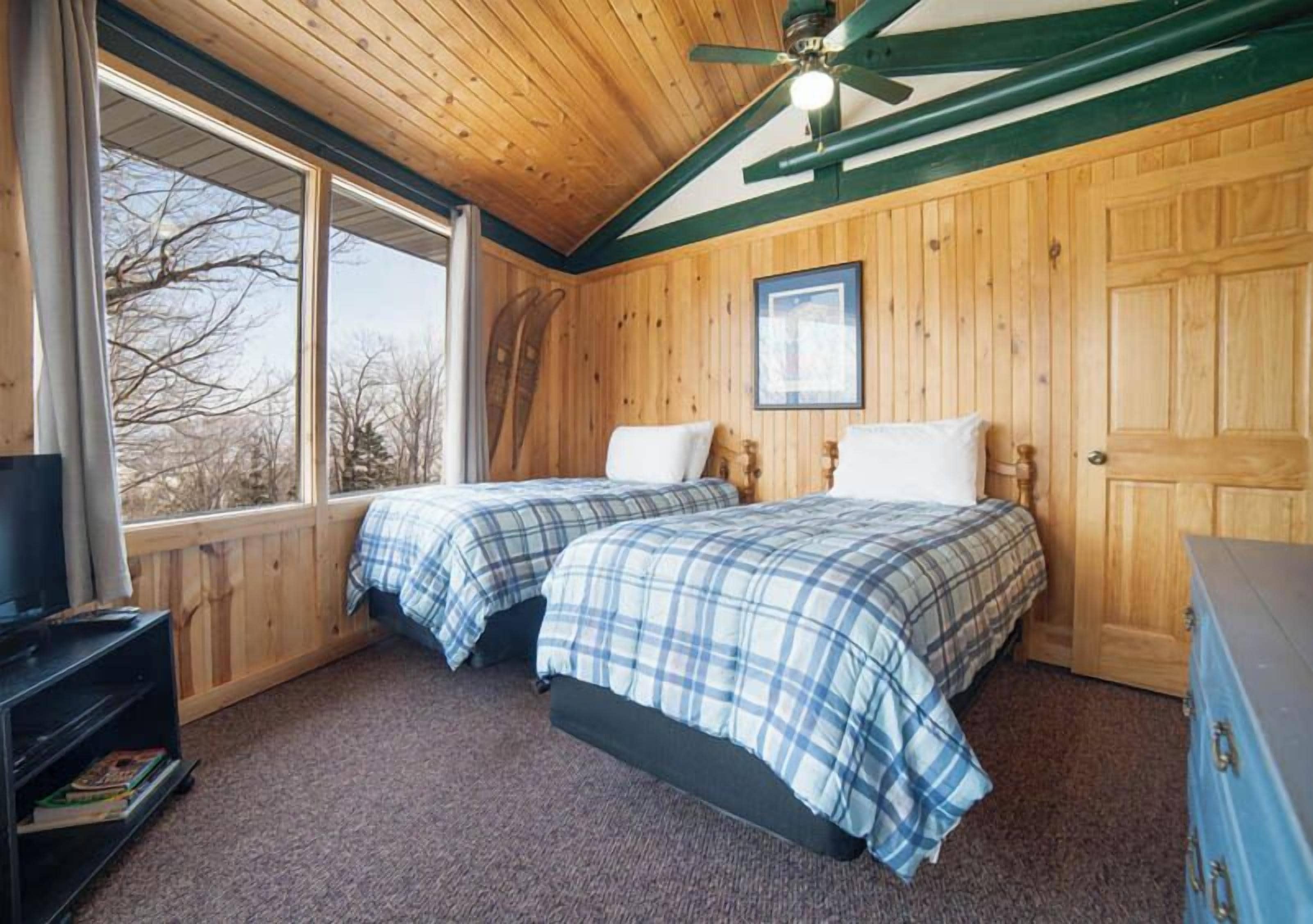 A room with two beds, wooden walls, a ceiling fan, and a window with a view of trees.
