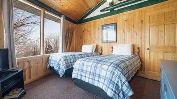 A room with two beds, wooden walls, a ceiling fan, and a window with a view of trees.