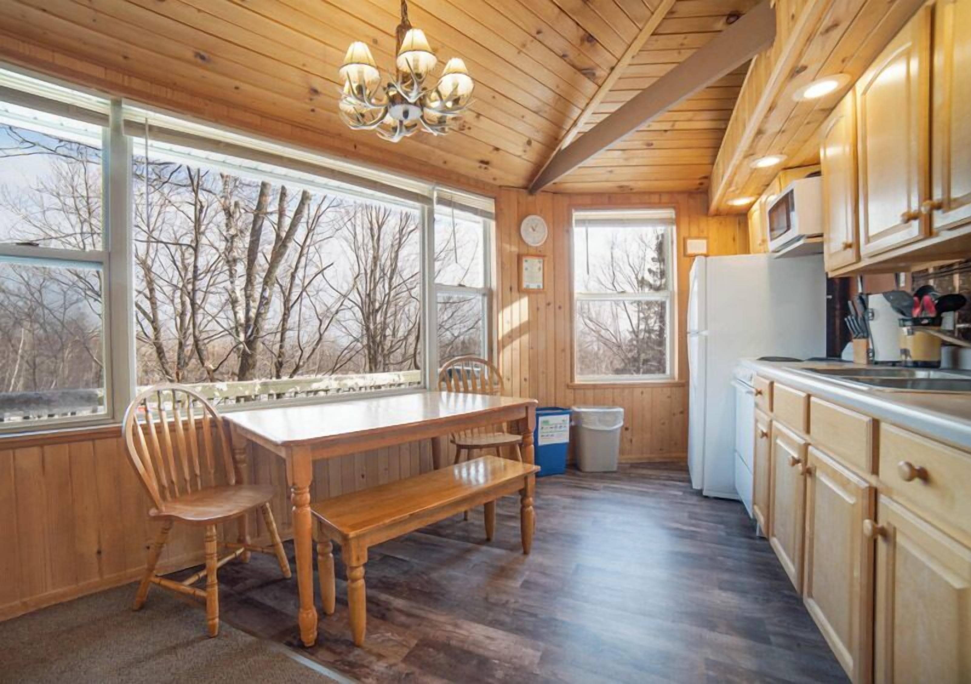 A wooden interior with a dining table and chairs, a kitchen with cabinets and appliances, and large windows with a view of bare trees.