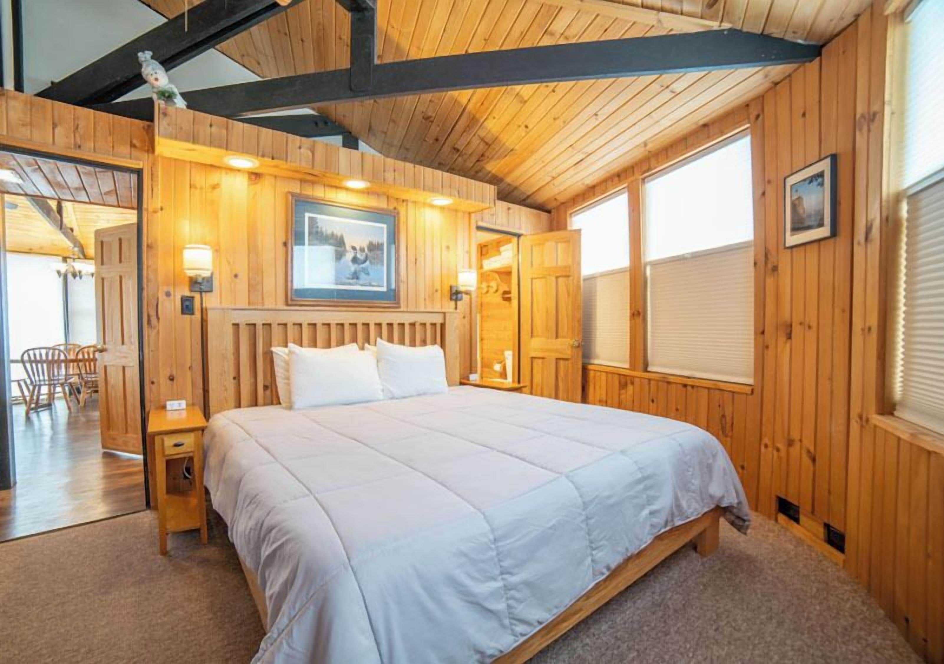 A wooden cabin bedroom with a bed, a nightstand, a framed picture on the wall, and a window.